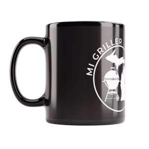 A glossy black ceramic coffee mug featuring high-contrast white text reading "I LIKE SMOKED BUTTS" next to the circular MI Griller kettle and Michigan silhouette logo, shown resting on a table.
